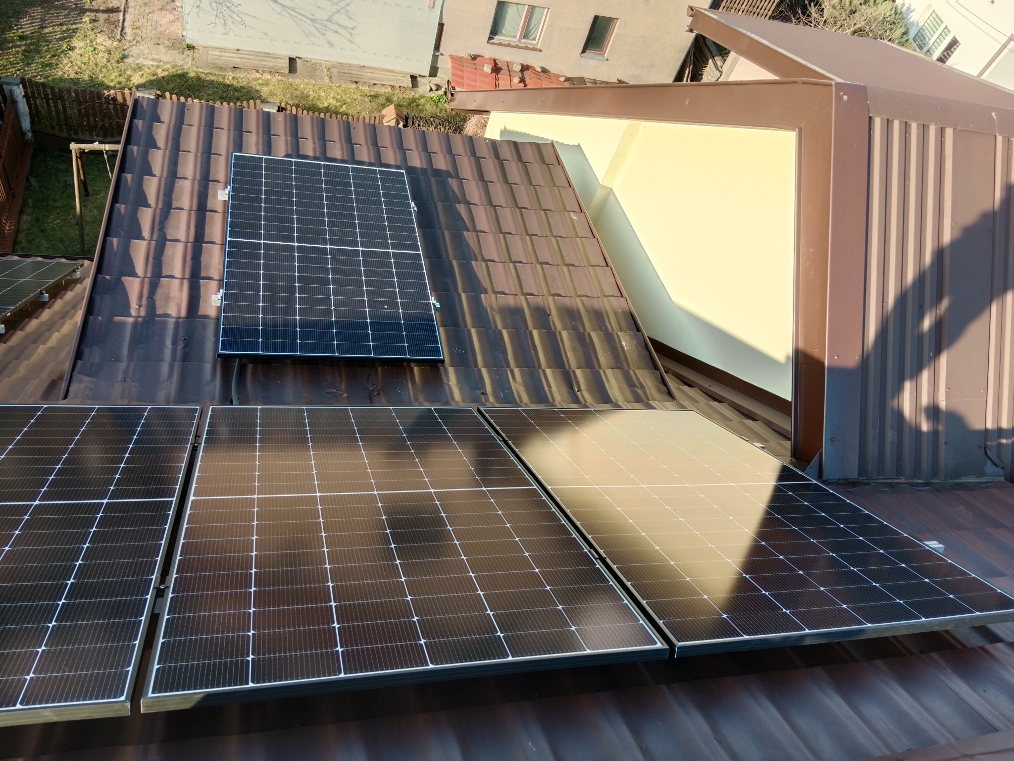 Panele fotowoltaiczne zamontowane na brązowym dachu z dachówki trapezowej, widok z góry, w tle budynki mieszkalne i drzewa.