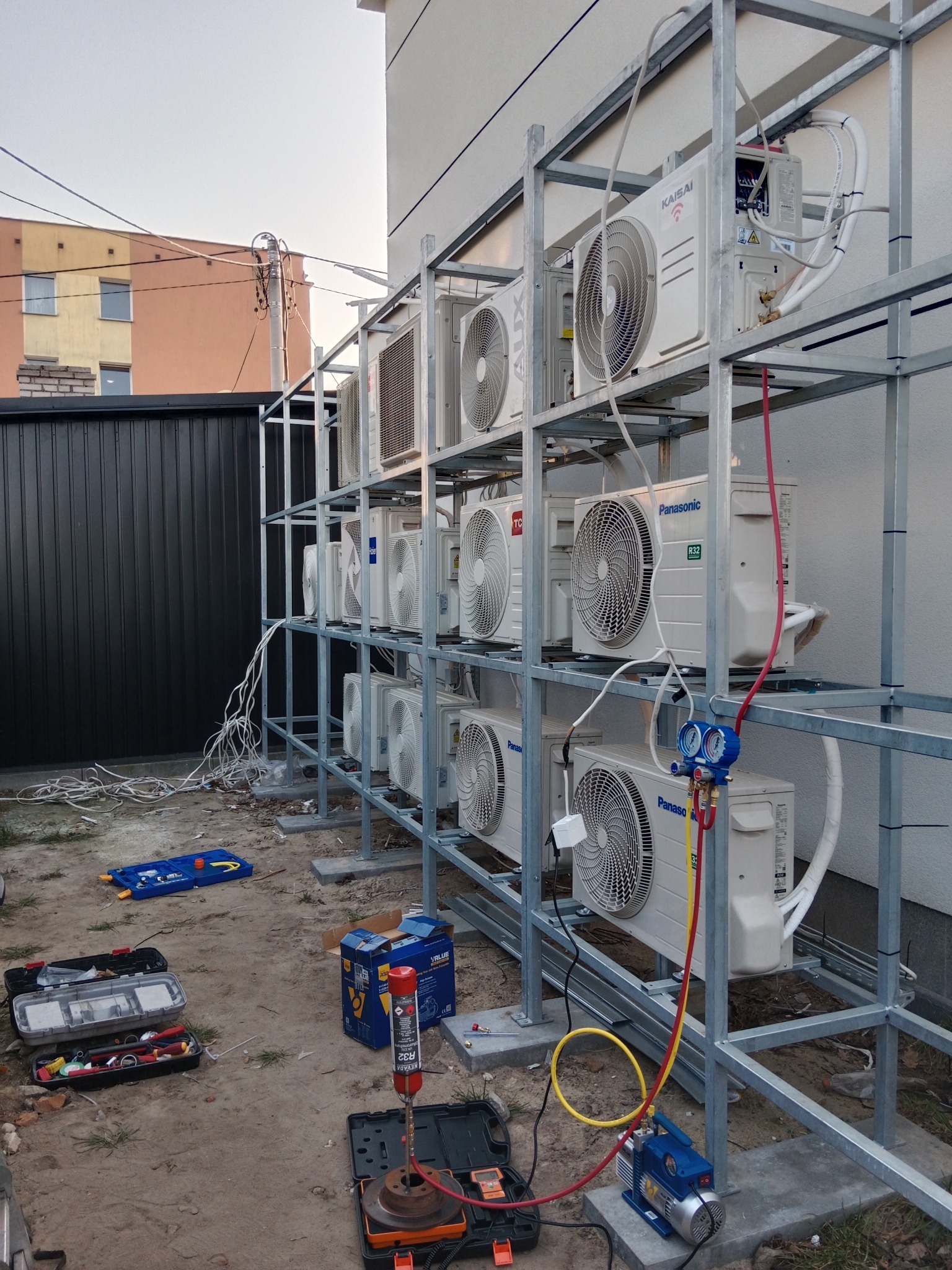 Rząd klimatyzatorów różnych marek (Kaisai, AUX, Panasonic, TC) zamontowanych na metalowym stelażu przy ścianie budynku, widoczne narzędzia i butla z gazem chłodniczym na ziemi, przewody i manometry...