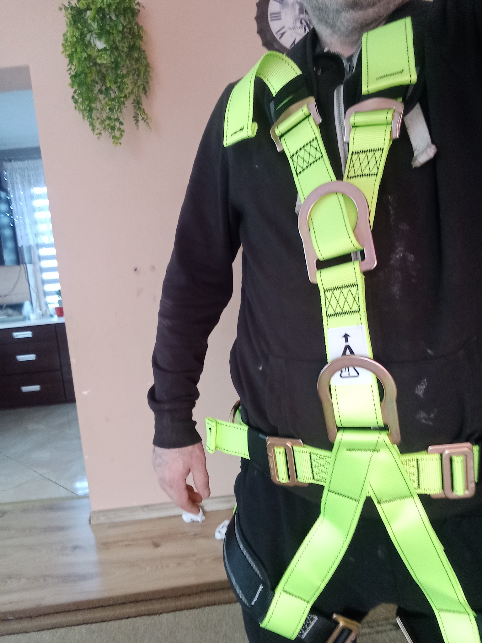 Mężczyzna w czarnej bluzie i neonowo-zielonej uprzęży bezpieczeństwa, z widocznym zegarem na ścianie w tle.