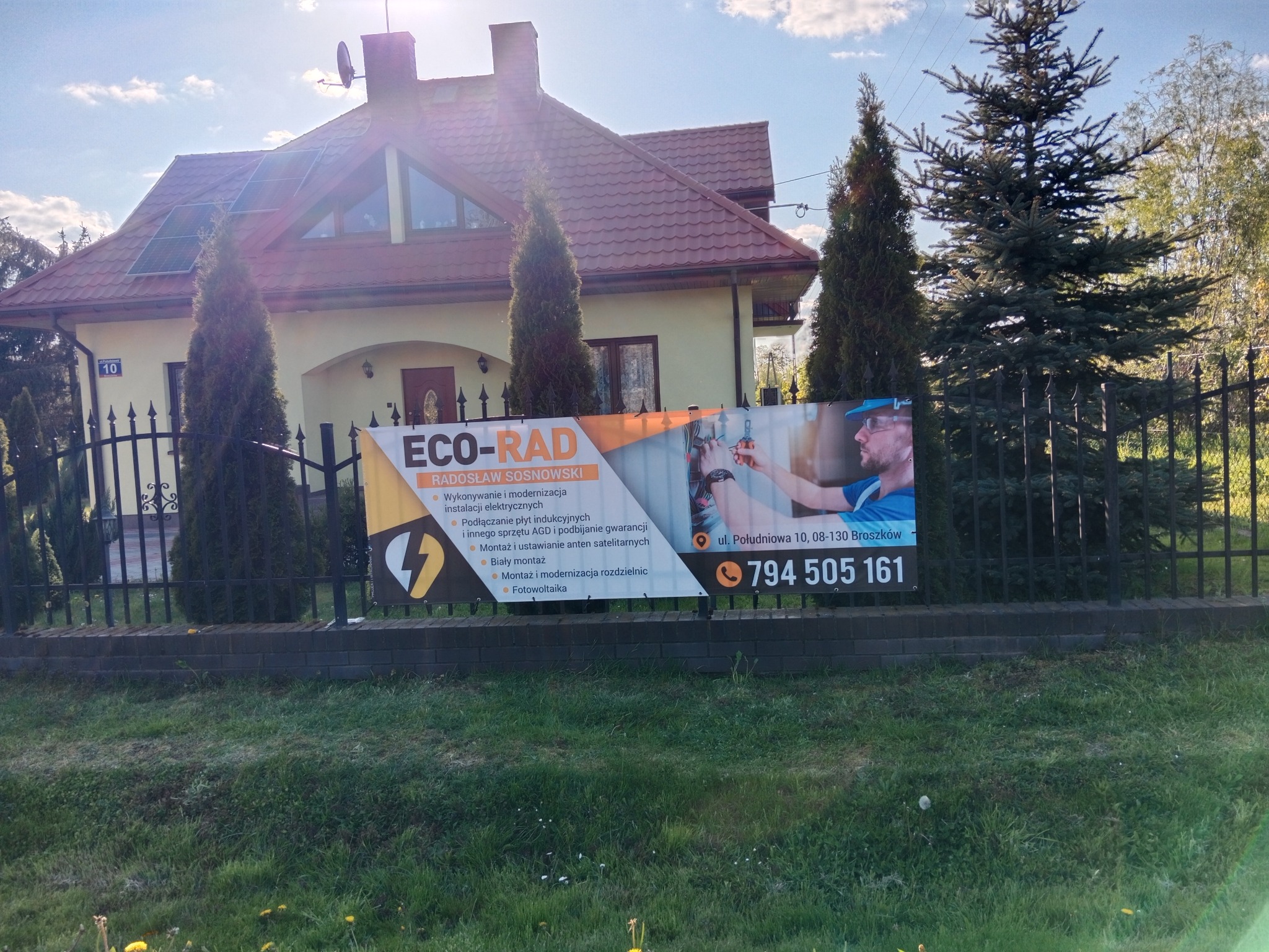 Baner firmy ECO-RAD na ogrodzeniu posesji z informacją o wykonywaniu i modernizacji instalacji elektrycznych, podłączaniu płyt indukcyjnych oraz montażu anten satelitarnych.