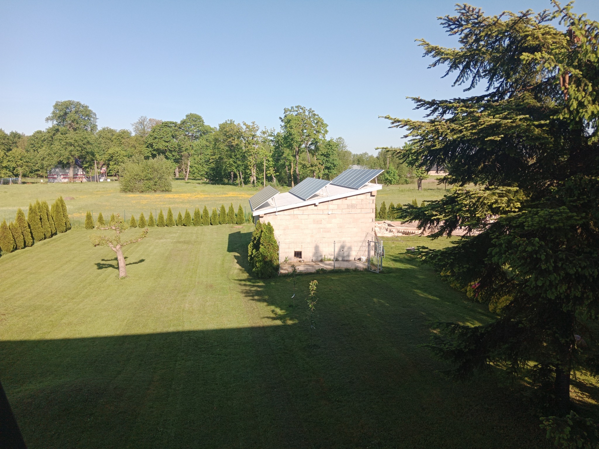 Niedokończony budynek gospodarczy z panelami słonecznymi na dachu, otoczony równo przystrzyżonym trawnikiem i rzędem tuj, widok z góry, cień w dolnej części kadru.