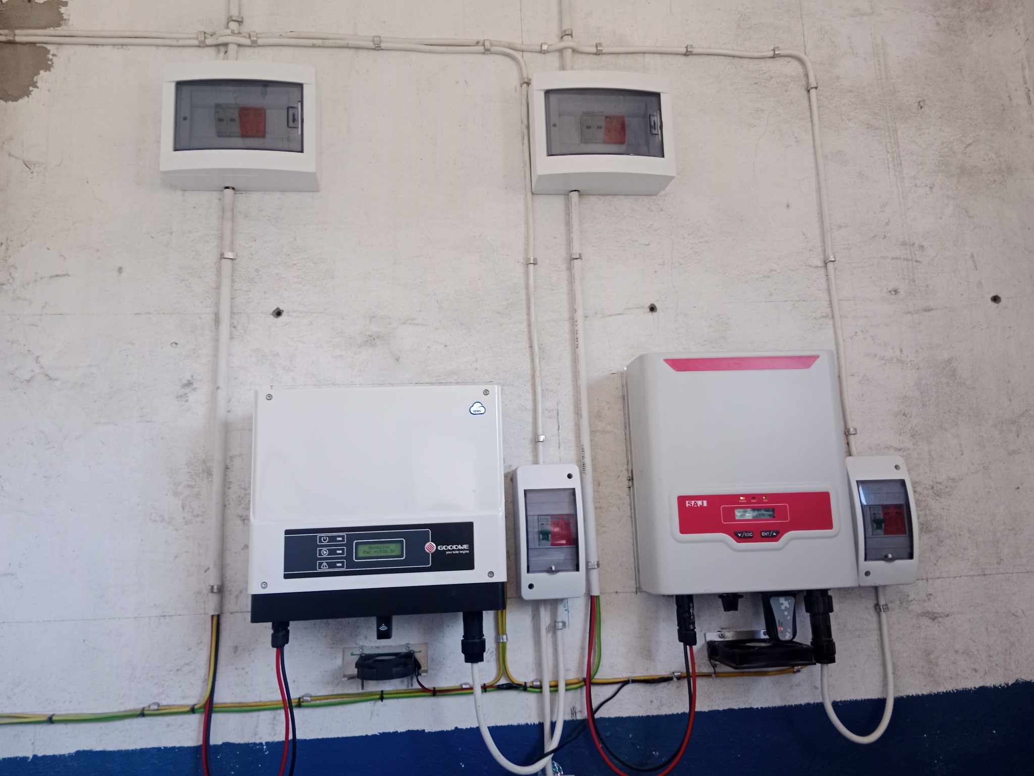 Dwa inwertery fotowoltaiczne, marki GoodWe i SAJ, zamontowane na ścianie z widocznymi przewodami i skrzynkami bezpiecznikowymi.
