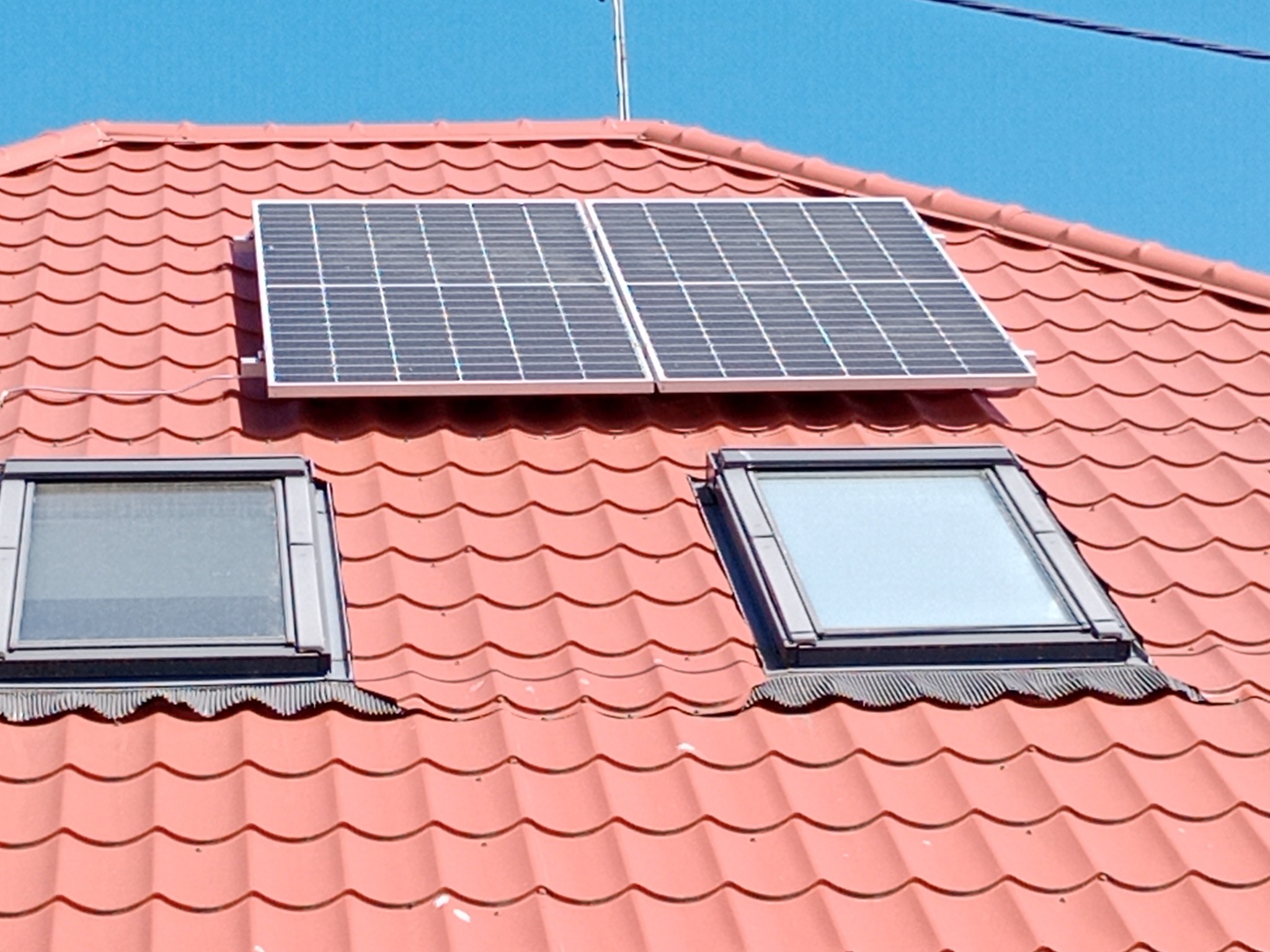 Dwa panele fotowoltaiczne zamontowane na czerwonym dachu z dachówką ceramiczną, obok dwóch okien dachowych.