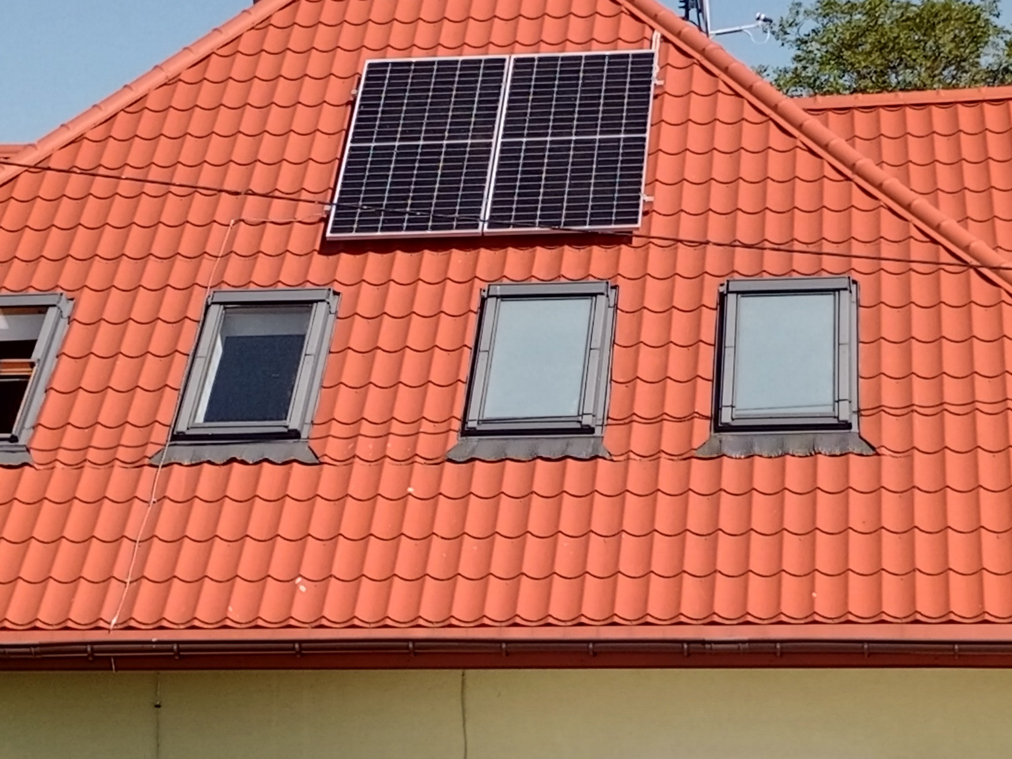 Dwa panele fotowoltaiczne zamontowane na pomarańczowym dachu z dachówki ceramicznej, widoczne trzy okna dachowe.