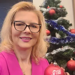 Uśmiechnięta blondynka w różowej marynarce, trzymająca czerwoną bombkę z logo Generali Group na tle choinki z napisem 'Wesołych Świąt'.