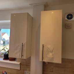 jakub surowiec - Nowo zamontowane, białe szafki kuchenne w Pogórzu, z folią ochronną na uchwytach, widoczne na tle okna z roślinami doniczkowymi i marmurowych płytek.