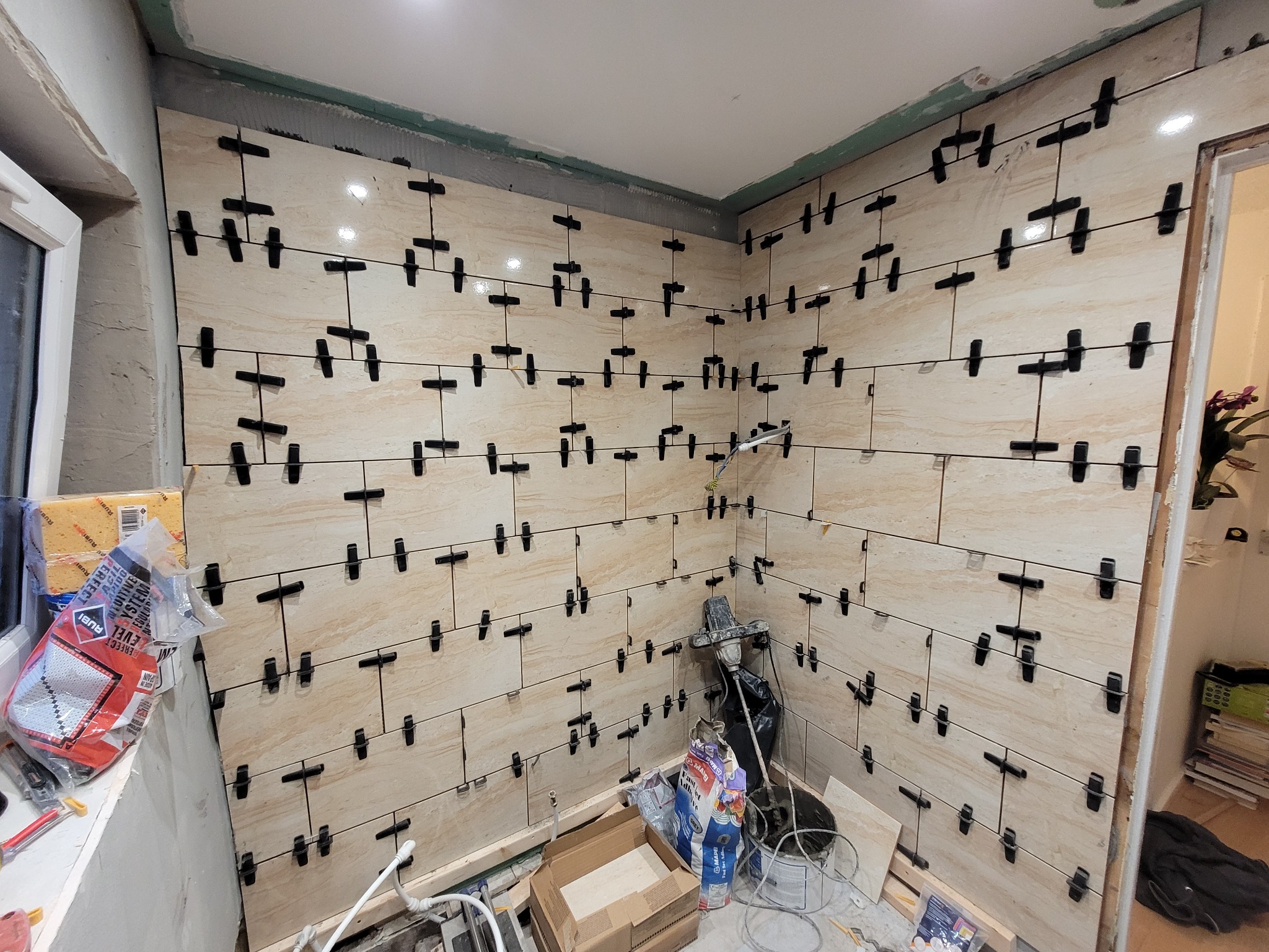 Ściana w trakcie układania beżowych płytek z użyciem klipsów poziomujących, widoczne narzędzia i materiały budowlane, w tle okno i fragment pomieszczenia.