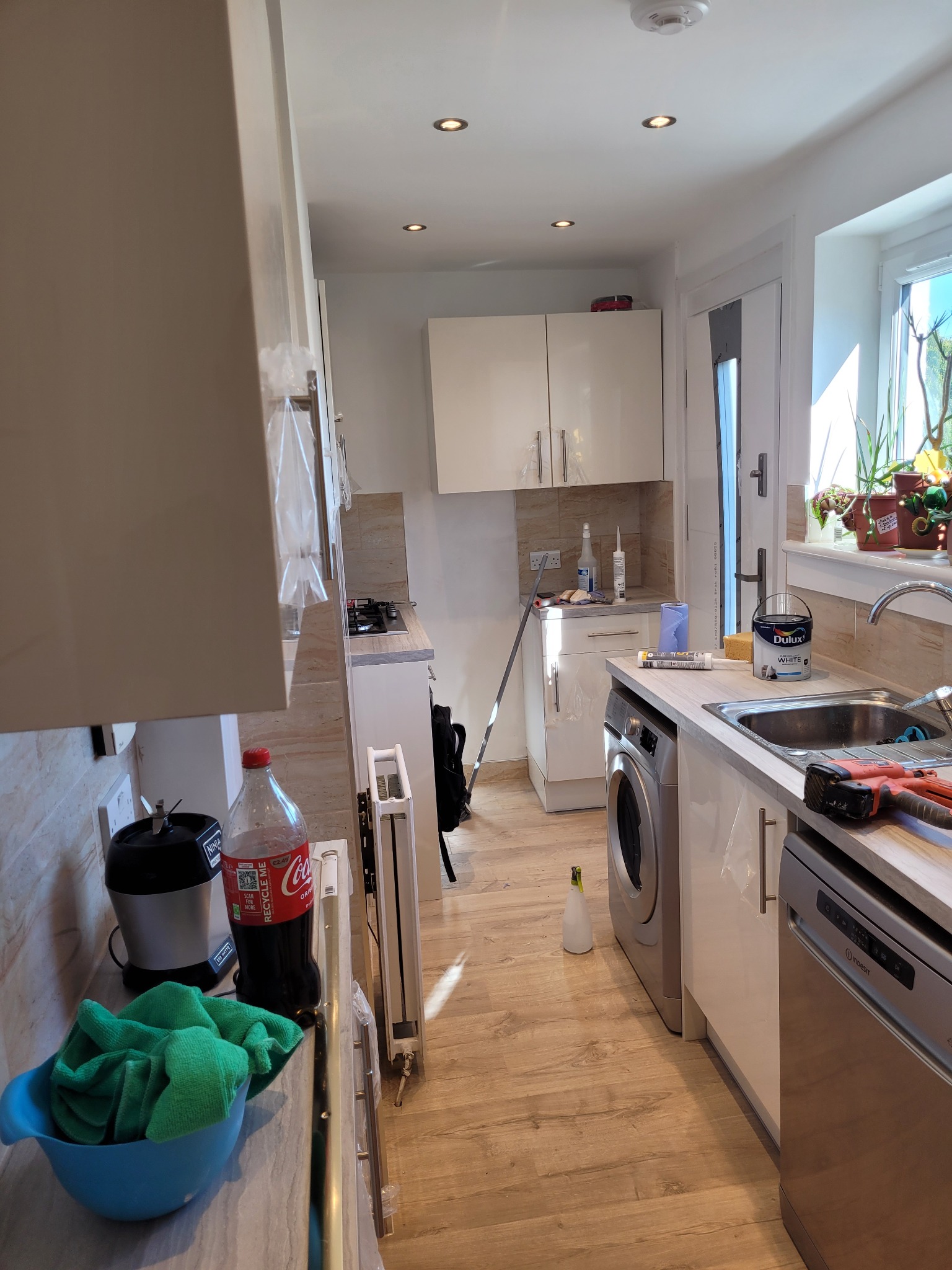 Wnętrze kuchni w trakcie prac, widoczny pojemnik z farbą Dulux, narzędzia, pralka i zmywarka, butelka Coca-Coli na blacie, podłoga wyłożona panelami, kuchnia w jasnych barwach z oświetleniem...