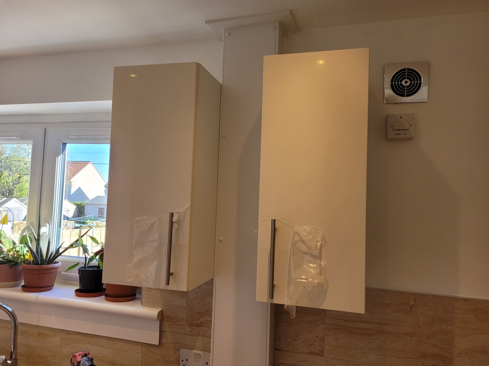 Nowo zamontowane, białe szafki kuchenne w Pogórzu, z folią ochronną na uchwytach, widoczne na tle okna z roślinami doniczkowymi i marmurowych płytek.