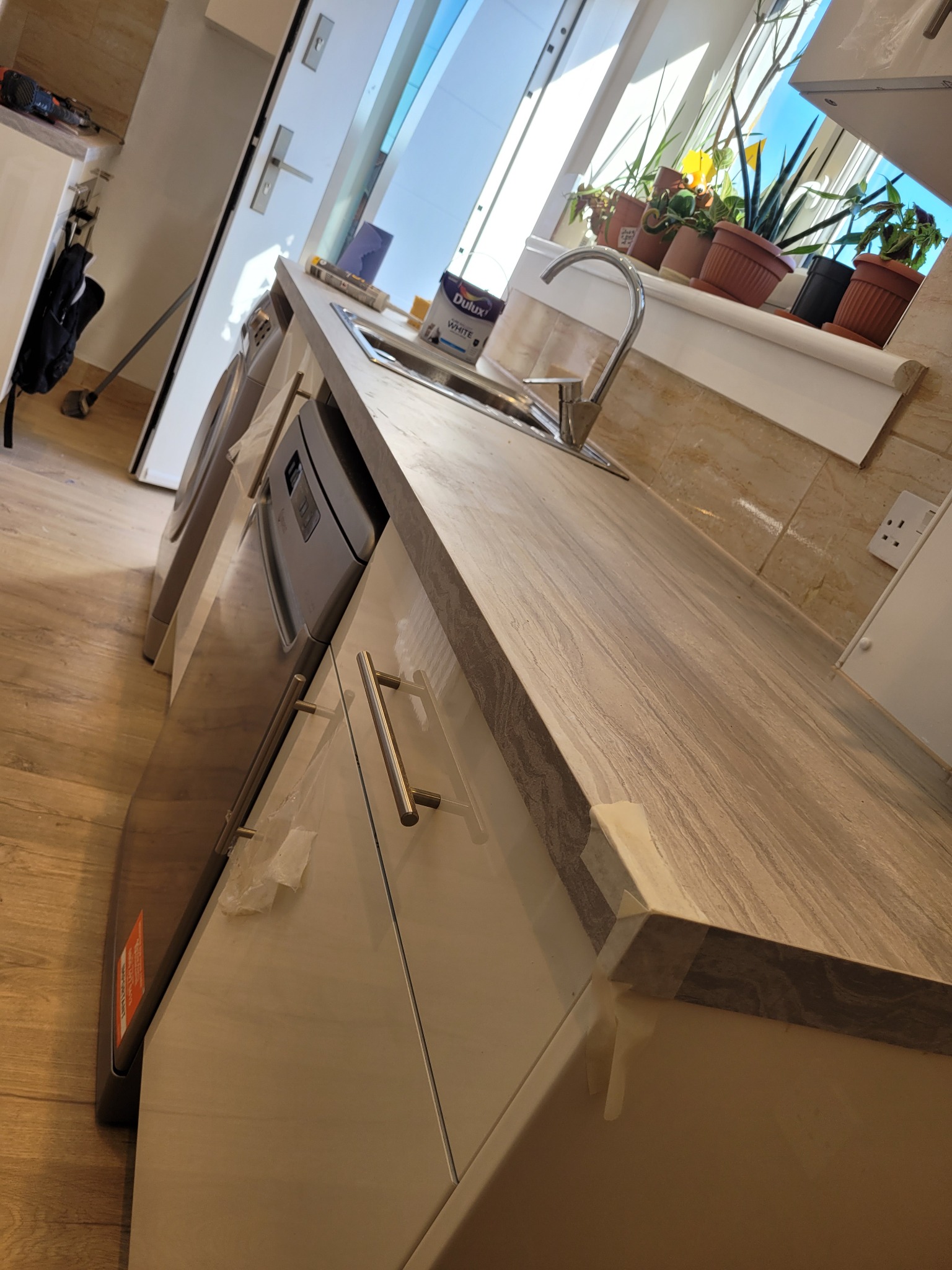 Nowa, jasna kuchnia z marmurowym blatem, chromowaną baterią i roślinami w doniczkach na parapecie, widoczna puszka farby Dulux White na blacie.