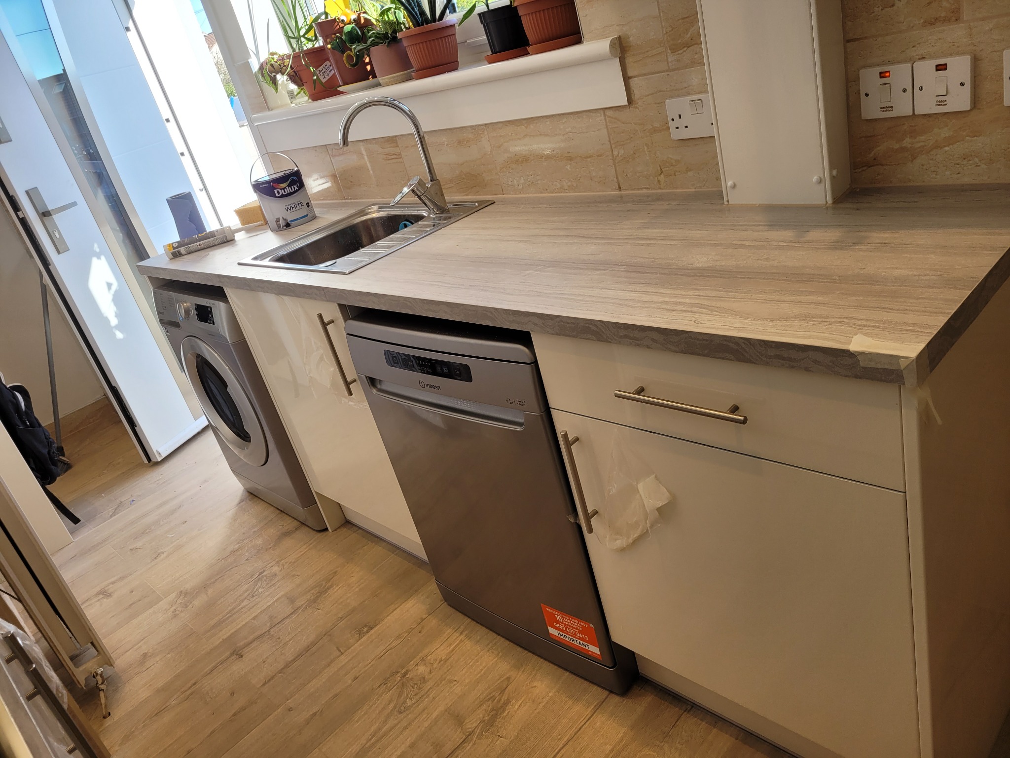 Nowoczesna kuchnia z drewnopodobnym blatem, zintegrowanym zlewozmywakiem, pralką i zmywarką w zabudowie, białe fronty szafek z poziomymi uchwytami, płytki imitujące kamień na ścianie, doniczki...