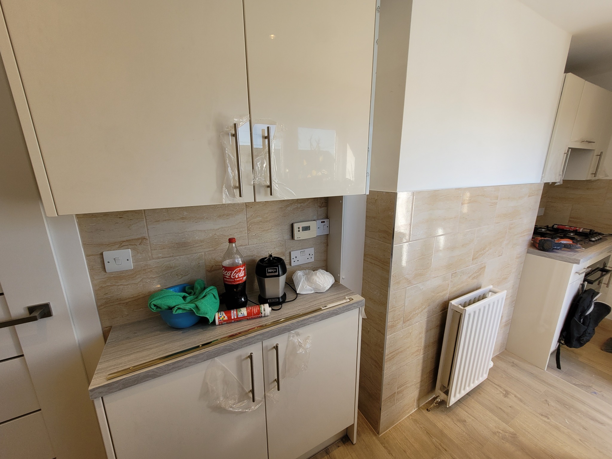 Jasna kuchnia w trakcie prac wykończeniowych: wiszące szafki z folią ochronną na uchwytach, beżowe płytki ścienne, grzejnik oraz akcesoria na blacie, w tym butelka Coca-Coli i blender.
