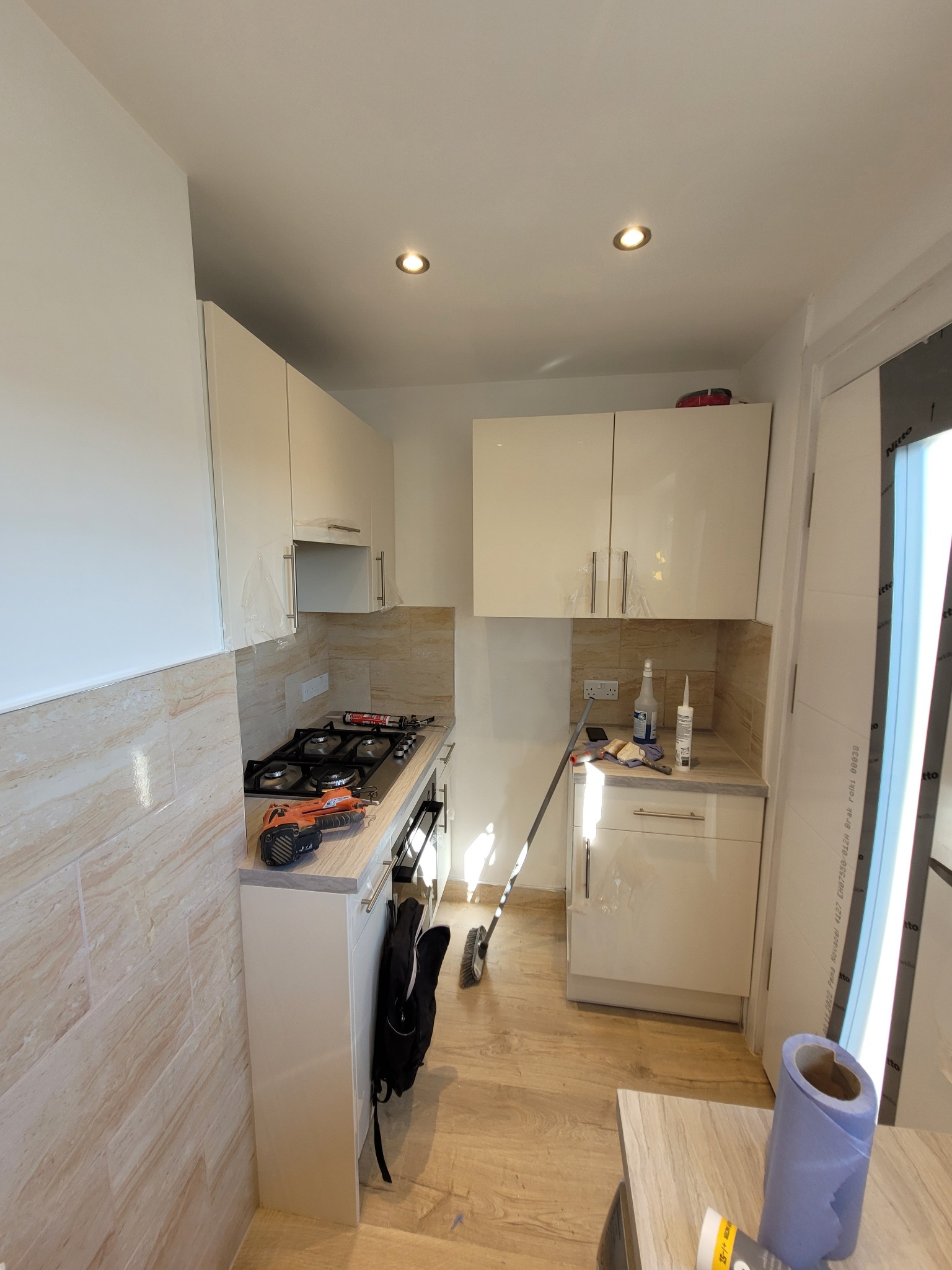 Wnętrze kuchni w trakcie prac wykończeniowych: widoczne białe szafki, beżowe płytki ścienne, narzędzia na blacie roboczym, częściowo zabezpieczone folią meble i niebieski ręcznik papierowy na stole.