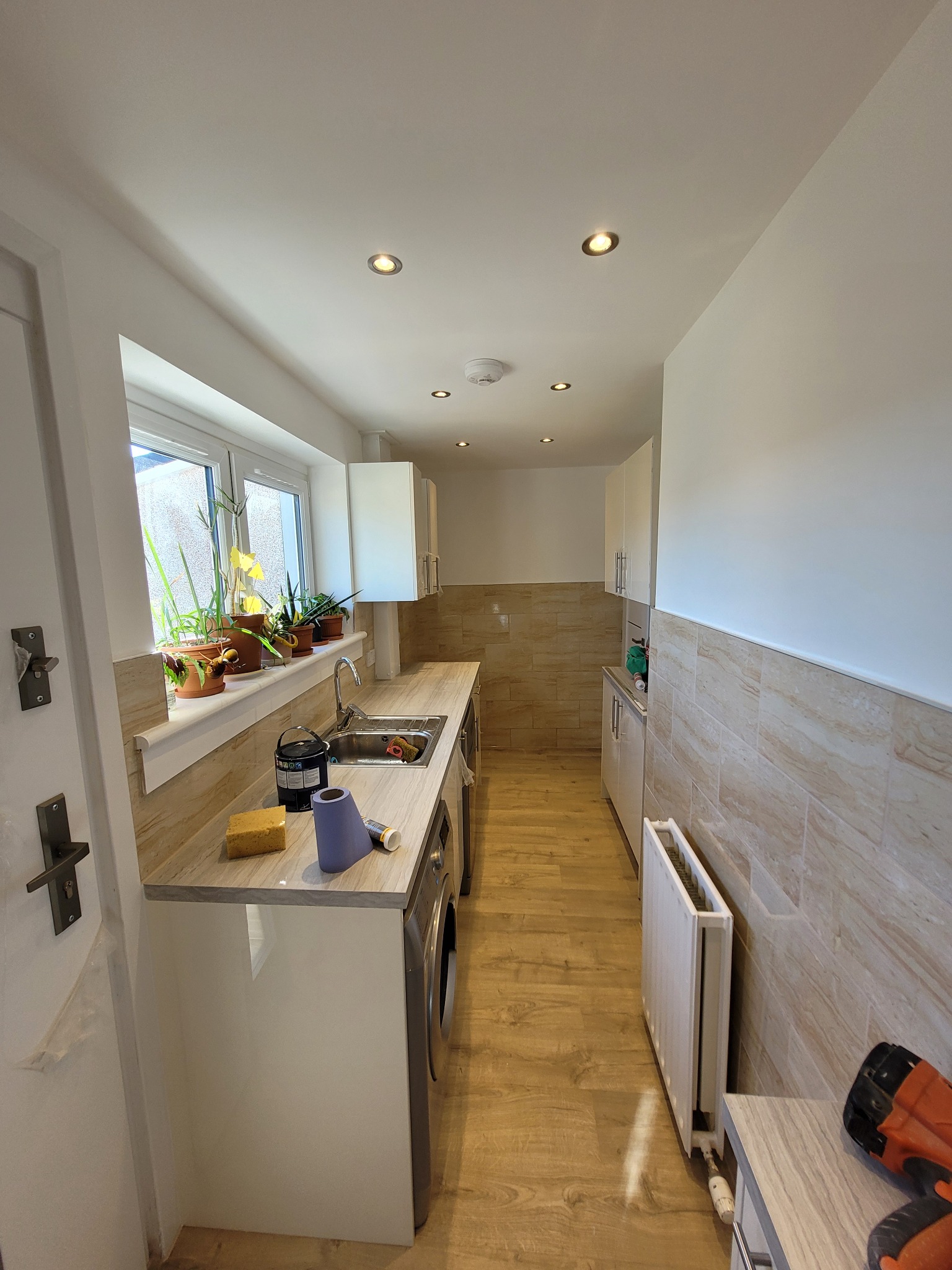 Wąska łazienka po remoncie z beżowymi płytkami imitującymi kamień, drewnianą podłogą, białymi szafkami i blatem, roślinami w doniczkach na parapecie i widoczną pralką.