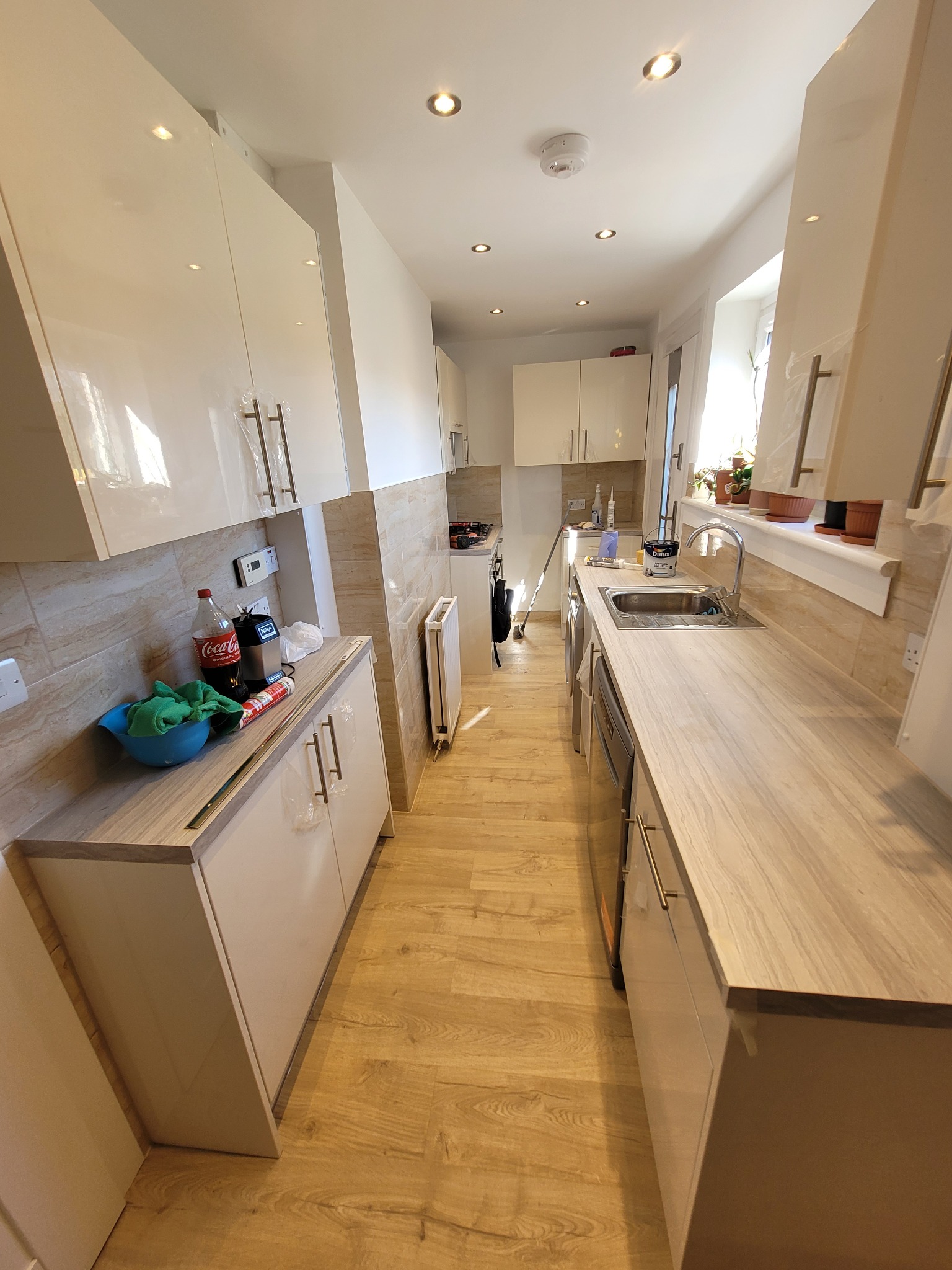 Wąska kuchnia z jasnymi, lakierowanymi szafkami, drewnopodobną podłogą i blatem, z widocznymi akcesoriami kuchennymi i narzędziami na blacie, sugerująca świeżo zakończony remont.