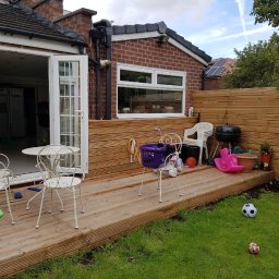 Olnada - Drewniany taras przydomowy z jasnymi meblami ogrodowymi, grillem i zabawkami, otoczony zielonym trawnikiem i drewnianym płotem.