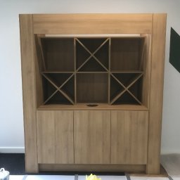 Olnada - Drewniana zabudowa meblowa z szafkami i otwartymi półkami z ukośnymi przegrodami, w jasnym odcieniu drewna, widoczna w trakcie montażu w pomieszczeniu biurowym z parkietem ułożonym we wzór jodełki.