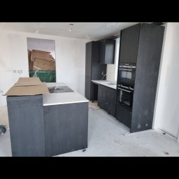 Olnada - Ciemna zabudowa kuchenna z wyspą i sprzętem AGD w trakcie montażu, widoczne kartony i niedokończone ściany.