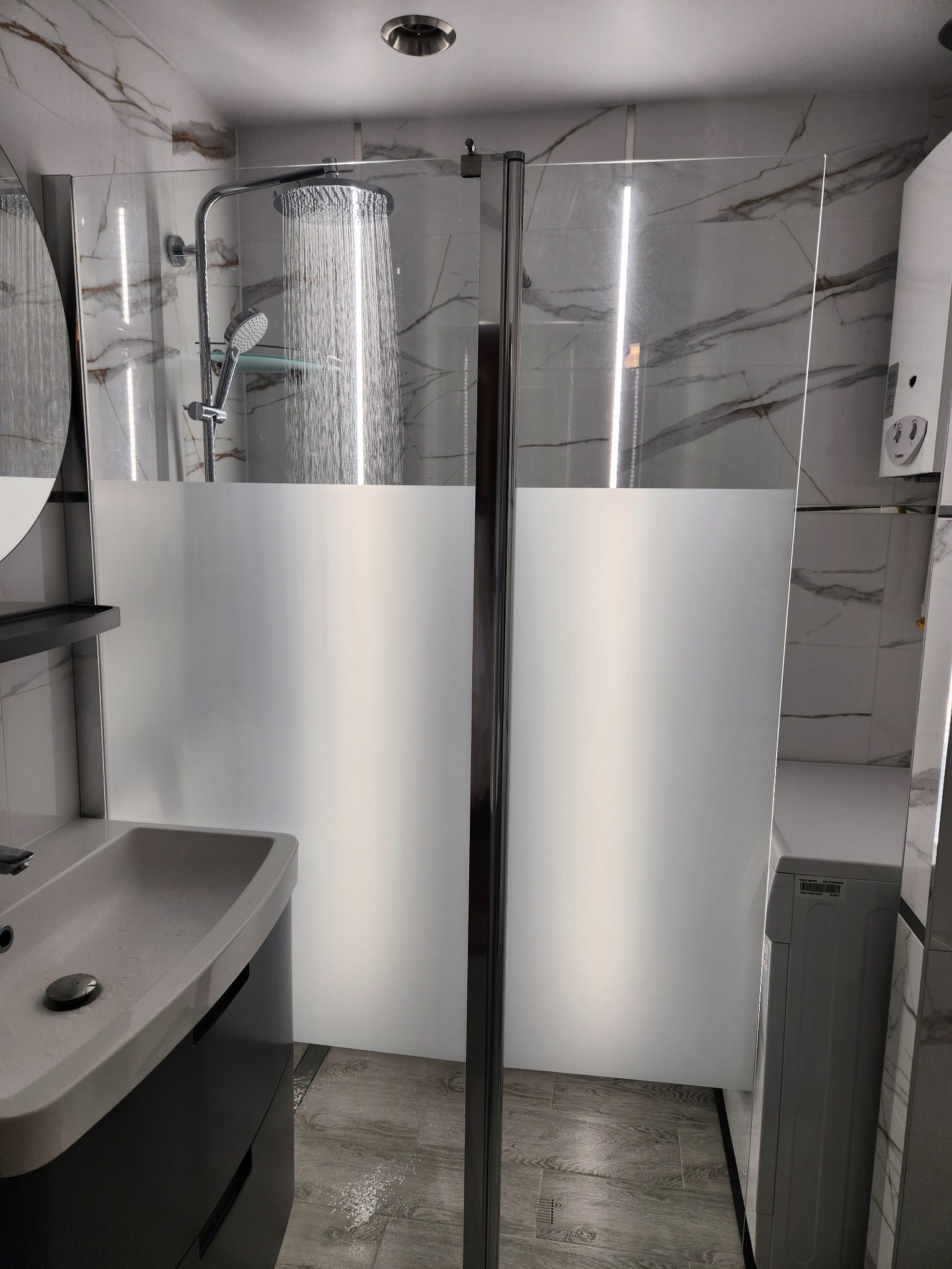 Nowoczesna kabina prysznicowa z deszczownicą i częściowo matową szybą, umywalka z szafką w kolorze grafitowym oraz marmurowe płytki na ścianach.