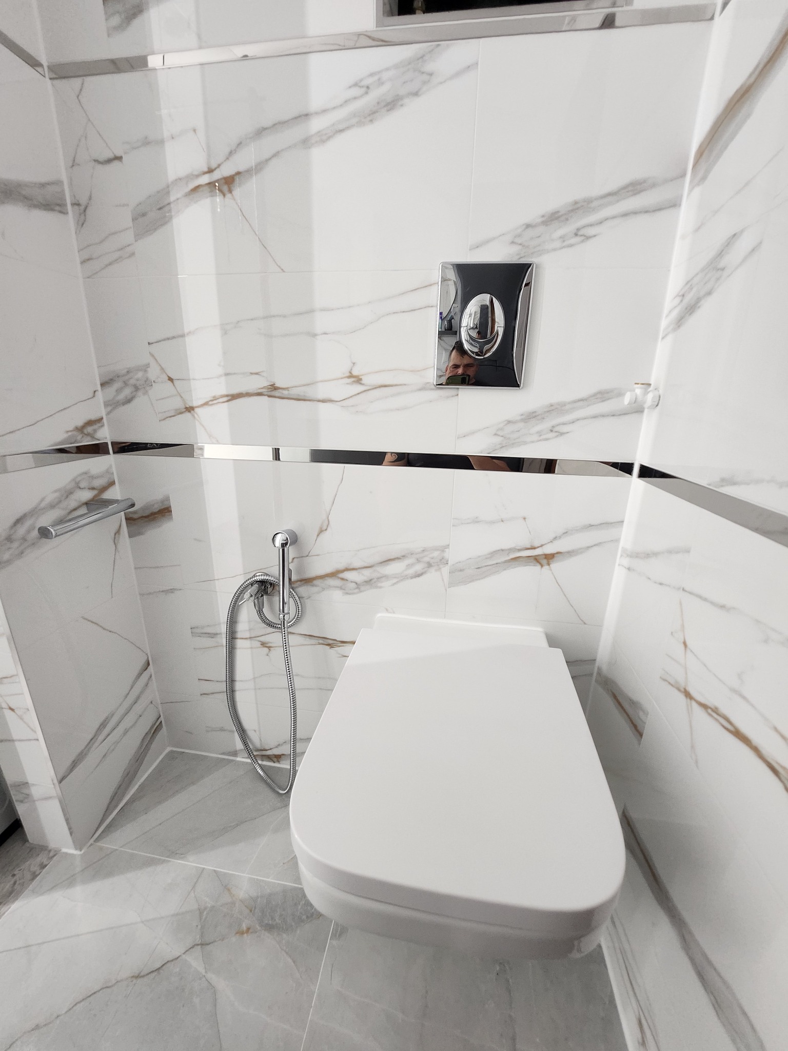 Nowoczesna toaleta podwieszana z bidetką i chromowanym przyciskiem spłukującym na tle białych płytek imitujących marmur z delikatnymi złotymi żyłami, wykończonych metalową listwą.