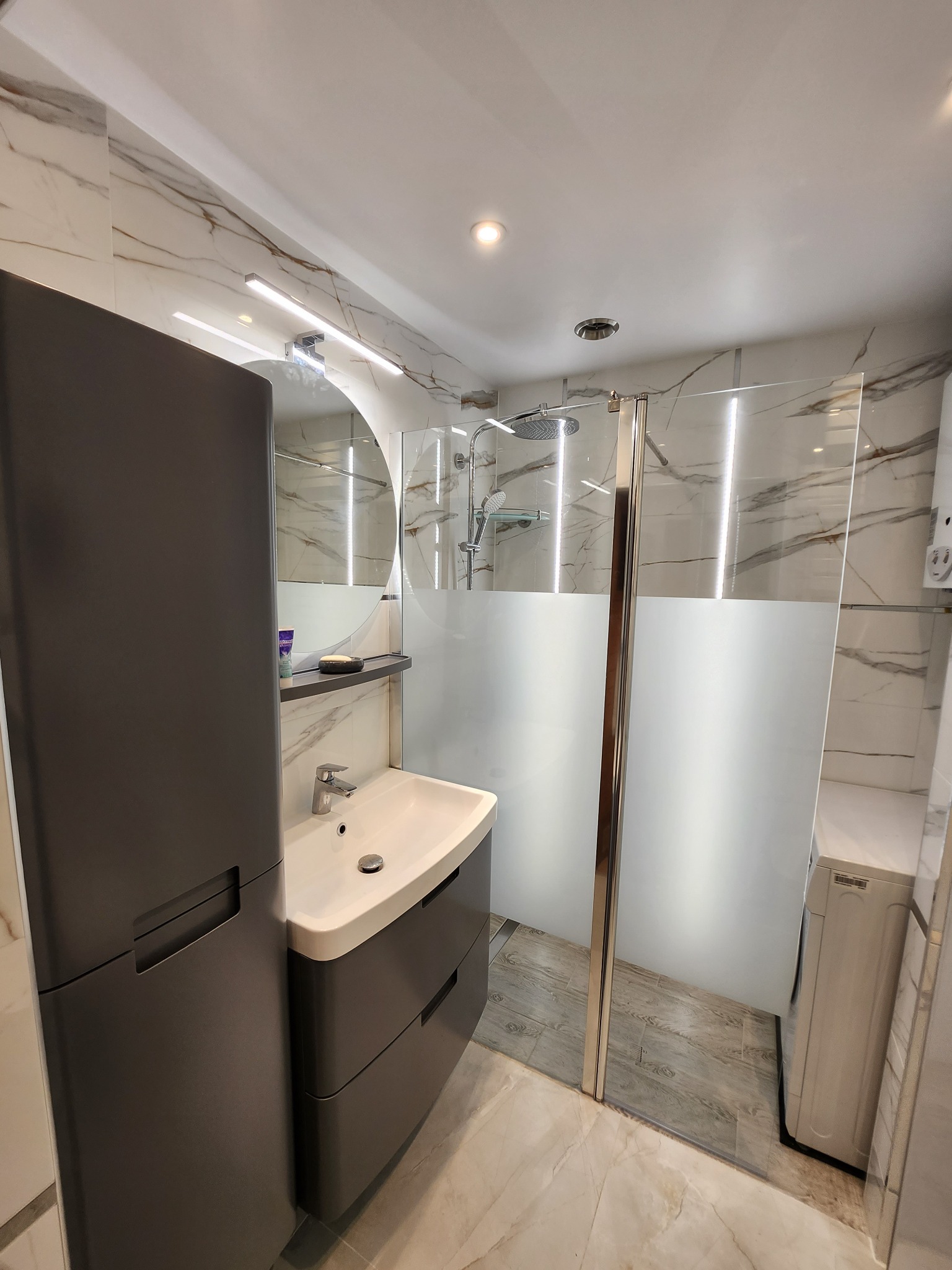 Nowoczesna łazienka z marmurowymi płytkami, szarą szafką umywalkową z szufladami, okrągłym lustrem z oświetleniem LED i kabiną prysznicową z częściowo matową szybą, w rogu widoczna pralka.