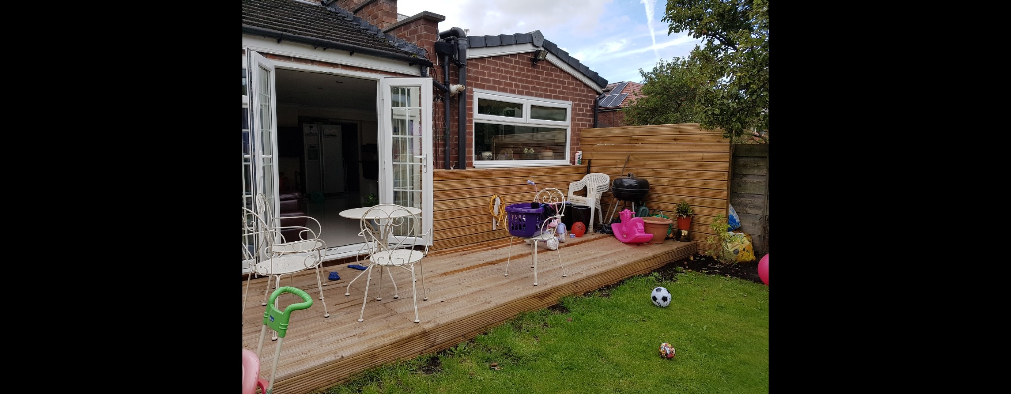 Drewniany taras przydomowy z jasnymi meblami ogrodowymi, grillem i zabawkami, otoczony zielonym trawnikiem i drewnianym płotem.