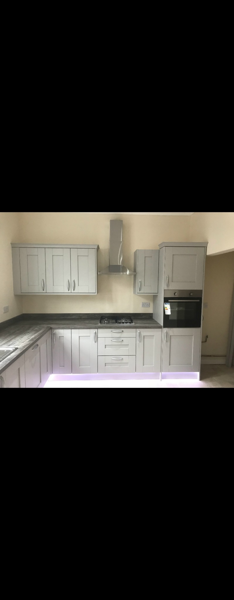 Jasnoszara, nowoczesna zabudowa kuchenna z oświetleniem LED pod szafkami, ciemny blat, płyta gazowa i piekarnik w zabudowie.