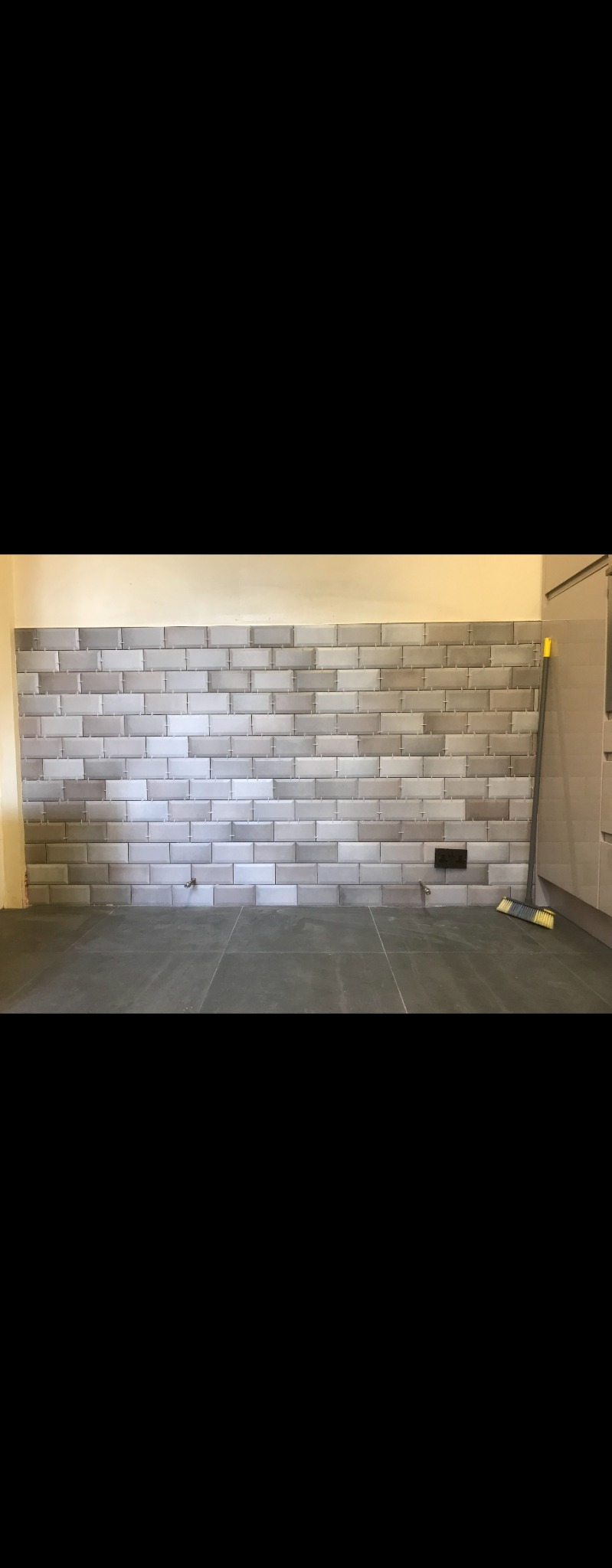 Ściana z ułożonych szarych płytek typu metro, z widocznymi fugami i wystającymi rurkami instalacji, na podłodze ciemne płytki, w tle fragment szafki i szczotka.