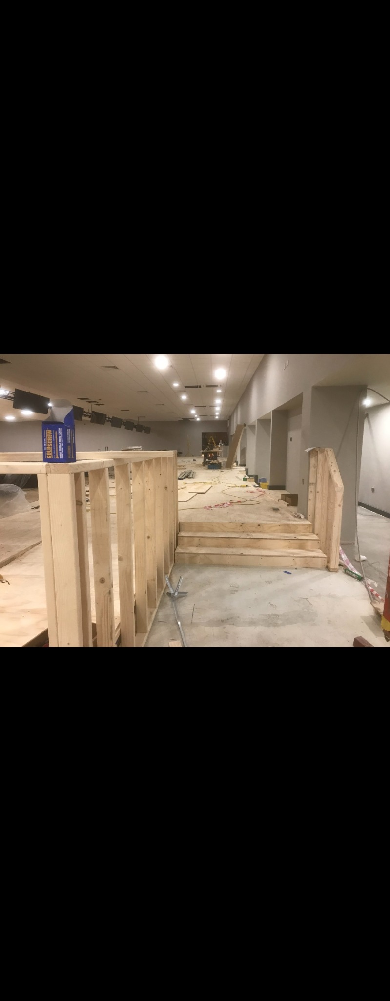 Wnętrze w trakcie prac budowlanych: surowy beton, drewniana konstrukcja schodów i ścianki działowej, widoczne narzędzia i materiały budowlane, oświetlenie sufitowe.