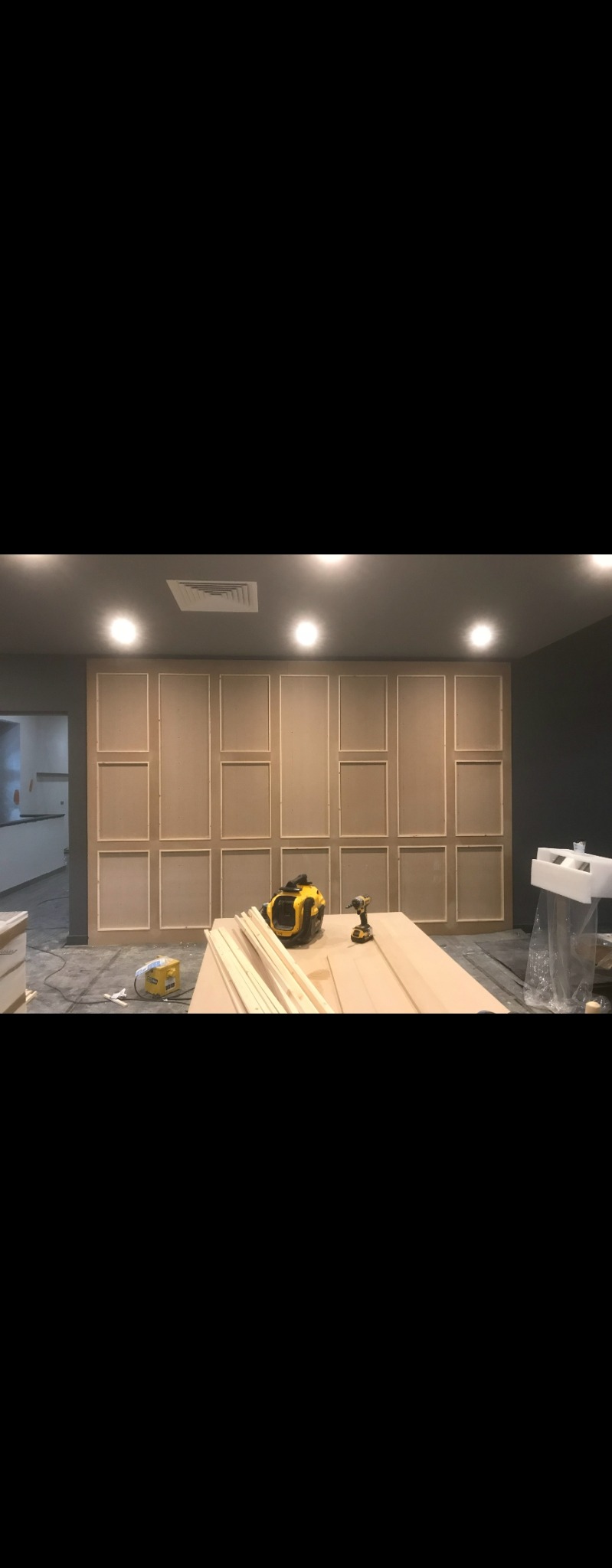 Ściana z paneli MDF w trakcie montażu, widoczne narzędzia: szlifierka i wkrętarka akumulatorowa, listwy przypodłogowe oraz płyta MDF na podłodze, surowy stan wykończenia.