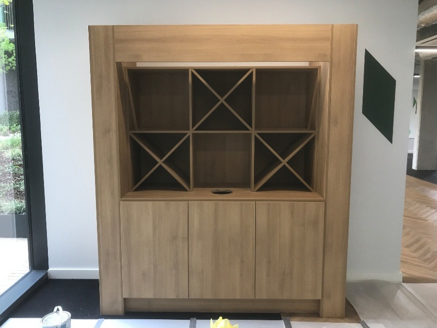 Drewniana zabudowa meblowa z szafkami i otwartymi półkami z ukośnymi przegrodami, w jasnym odcieniu drewna, widoczna w trakcie montażu w pomieszczeniu biurowym z parkietem ułożonym we wzór jodełki.