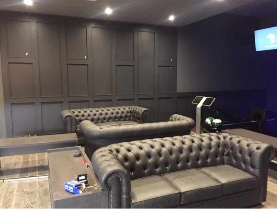 Eleganckie wnętrze w odcieniach szarości z panelami ściennymi, skórzanymi sofami Chesterfield i elementami wyposażenia siłowni.