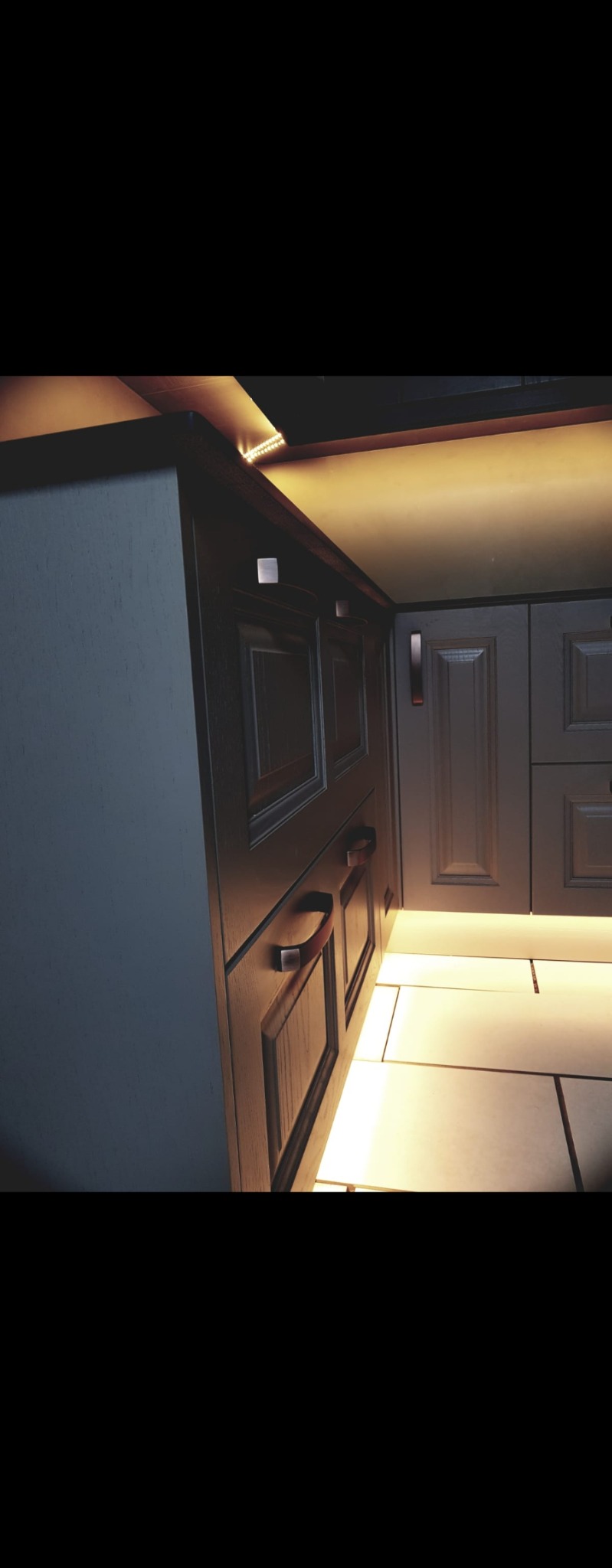 Szczegółowe ujęcie dolnych szafek kuchennych z oświetleniem LED pod blatem i nad podłogą, z widocznymi uchwytami i płytkami podłogowymi.