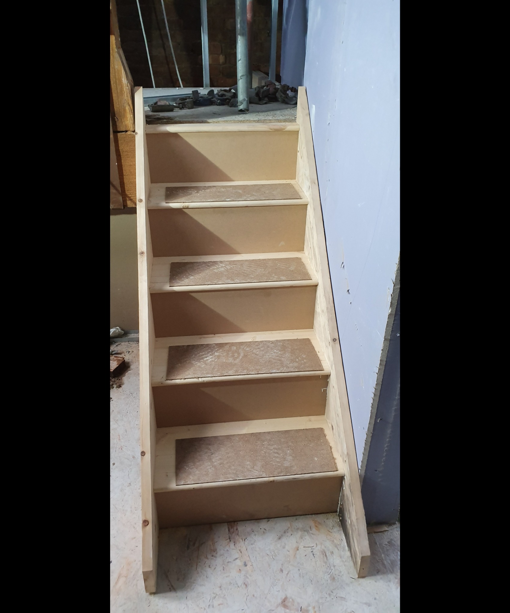 Surowe drewniane schody z płytą MDF na stopniach, widoczne w trakcie budowy, z elementami konstrukcyjnymi w tle i fragmentem ściany z płyt kartonowo-gipsowych.