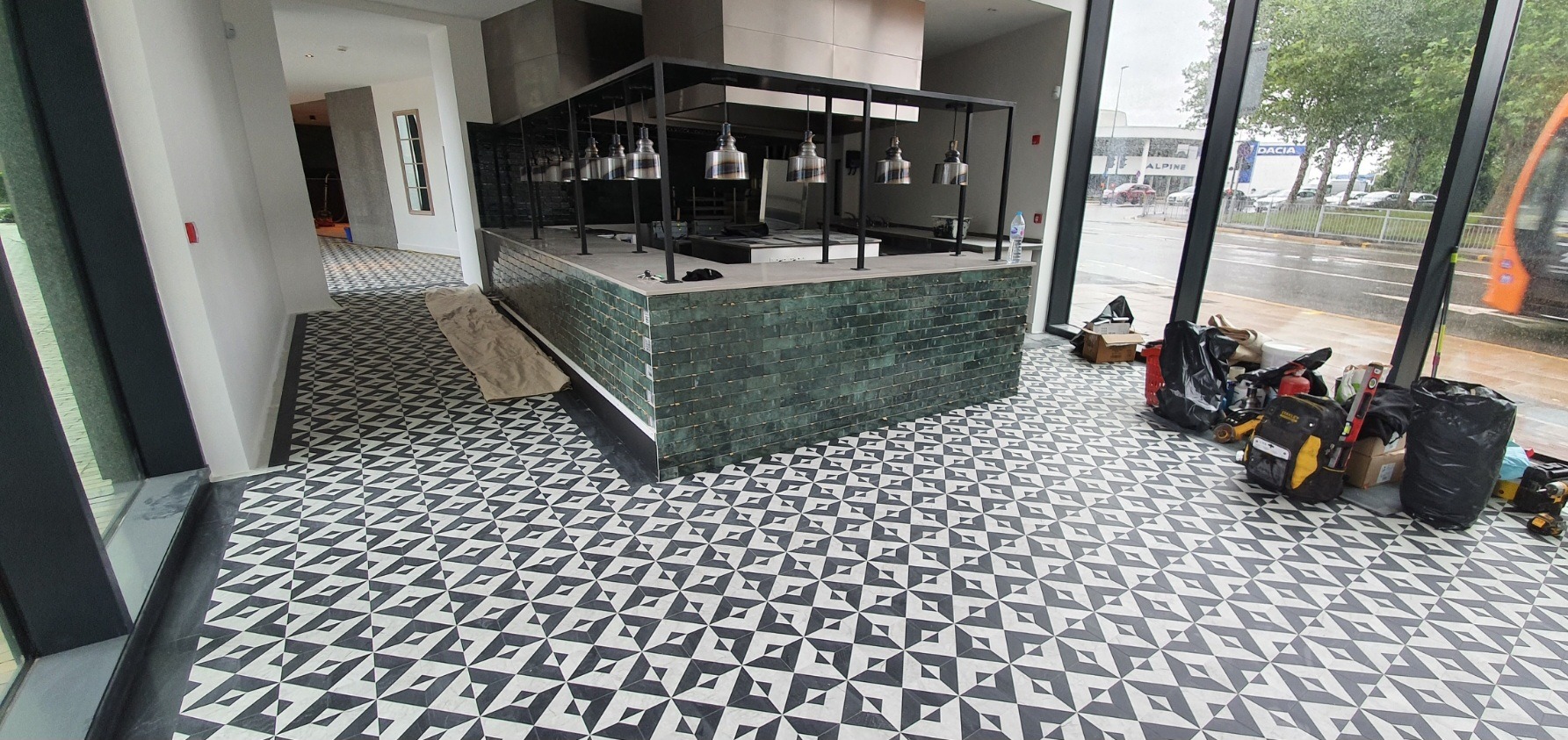 Nowa, wzorzysta posadzka w lokalu komercyjnym, z widocznym fragmentem baru wykończonego zieloną płytką oraz narzędziami i workami budowlanymi obok okna.