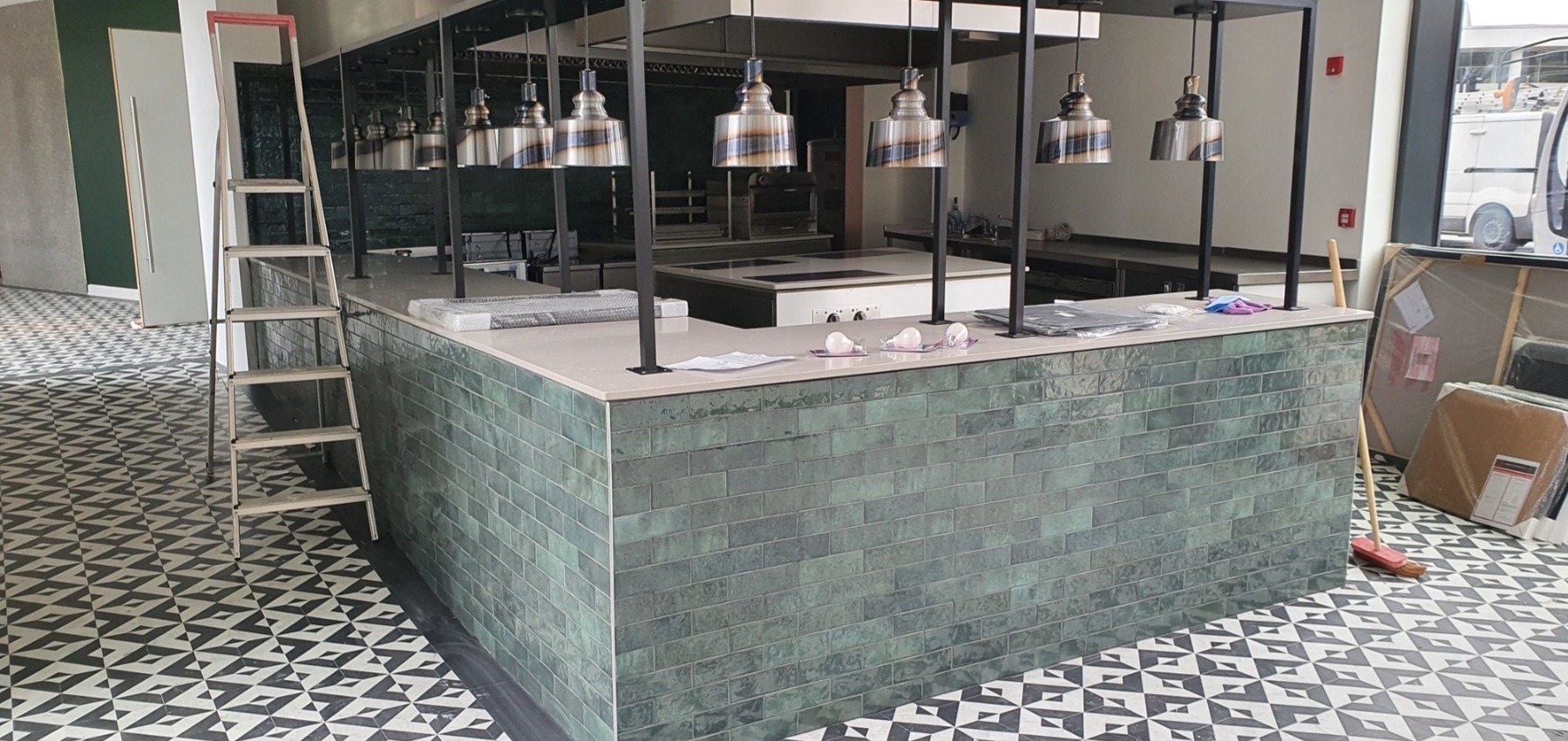 Bar z zielonej cegły z metalowymi lampami wiszącymi, drabina oparta o ścianę, podłoga z geometrycznym wzorem, w tle zaplecze kuchenne w trakcie wykańczania.