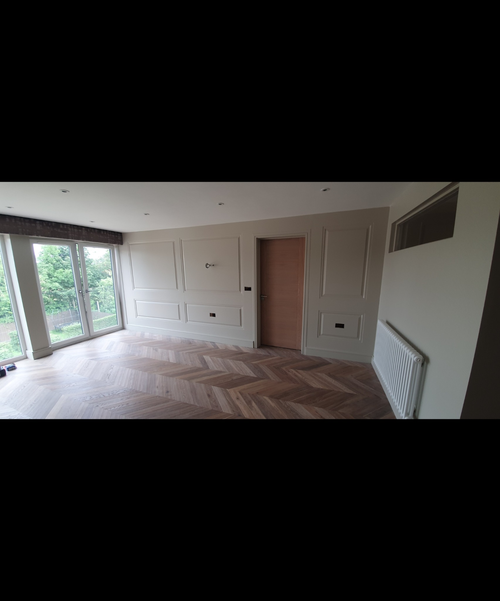 Jasne wnętrze z parkietem ułożonym w jodełkę, panelami ściennymi i grzejnikiem, widok na zieleń za oknem.