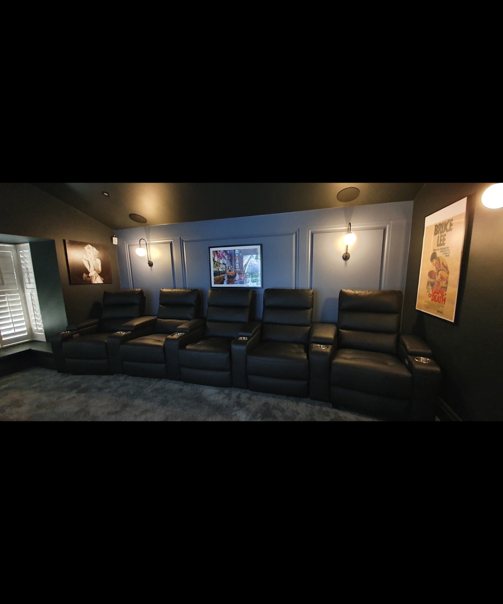Kino domowe z pięcioma czarnymi, skórzanymi fotelami z uchwytami na napoje, podświetlonymi kinkietami i dekoracjami ściennymi, w tym plakatem Bruce Lee.