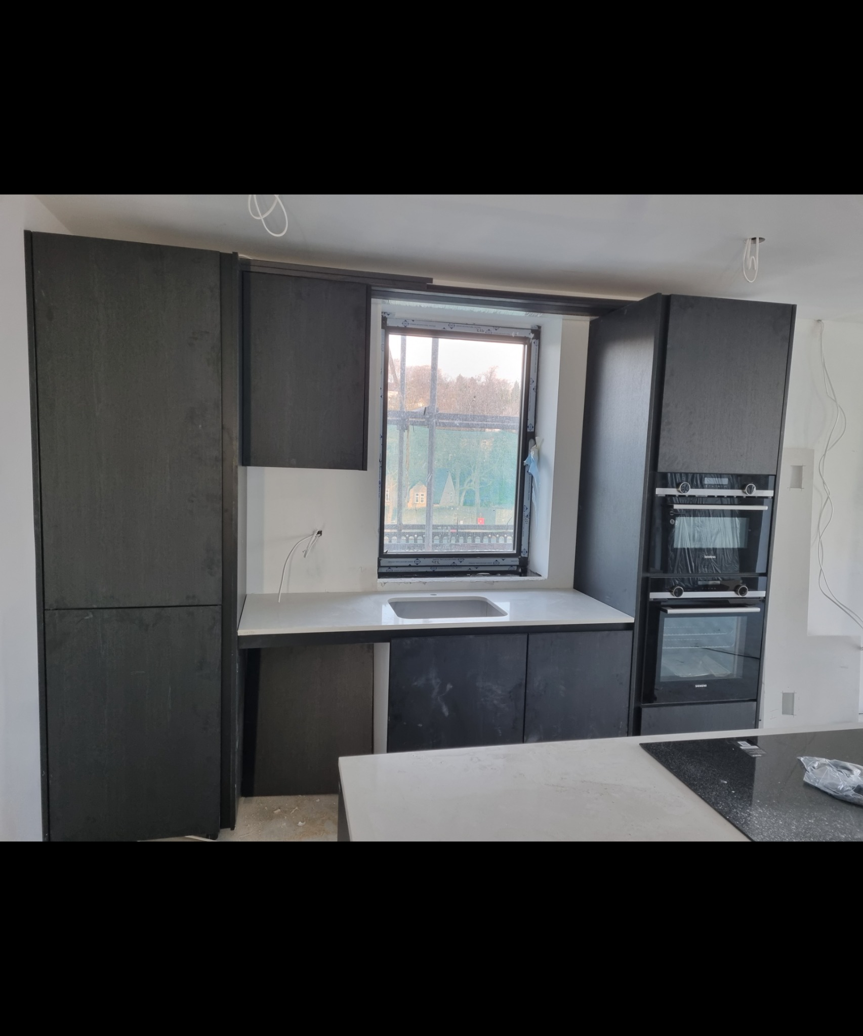 Ciemne, nowoczesne szafki kuchenne z białym blatem i zlewem pod oknem, piekarniki w zabudowie, instalacja elektryczna widoczna na ścianie.