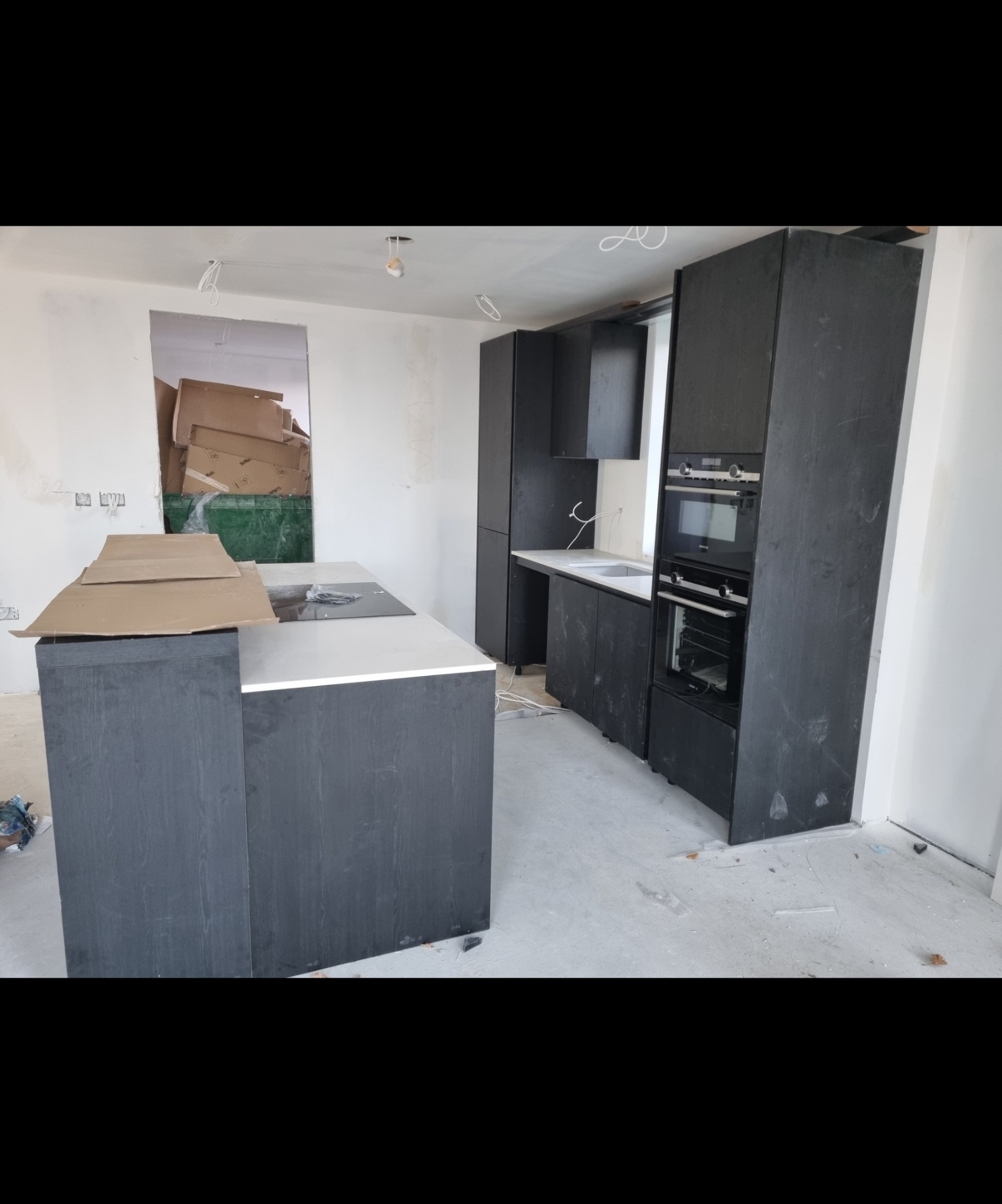 Ciemna zabudowa kuchenna z wyspą i sprzętem AGD w trakcie montażu, widoczne kartony i niedokończone ściany.