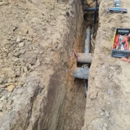 Głęboki wykop w ziemi z ułożonymi rurami, widoczna skrzynka z narzędziami i poziomica laserowa na krawędzi wykopu.