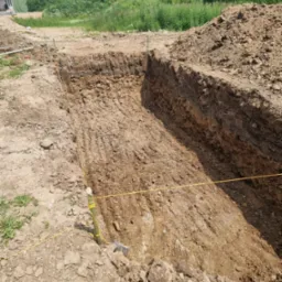 Wykop pod fundamenty z widocznymi warstwami gleby, wyznaczony sznurkiem i palikami, w tle zieleń i fragment domu.