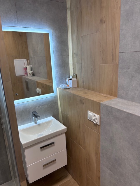 Nowoczesna łazienka z podświetlanym lustrem, umywalką z szafką i płytkami imitującymi drewno. Widoczny kontakt na ścianie. Minimalistyczny design.