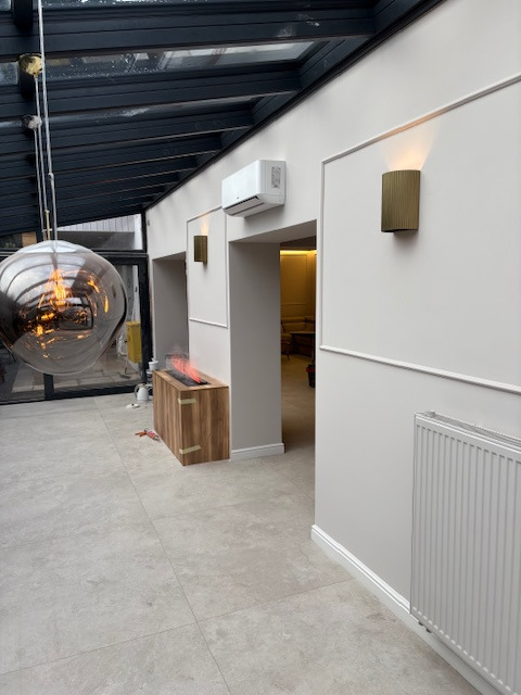 Jasne wnętrze z klimatyzacją, ozdobnymi listwami ściennymi i nowoczesnym oświetleniem. Widoczny grzejnik, podłoga z płytek i przeszklony sufit.