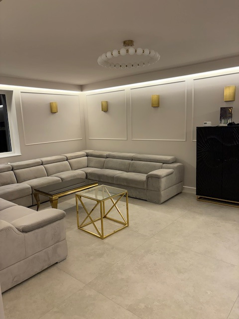 Elegancki salon z narożnikiem w odcieniach szarości, stolikami kawowymi ze złotymi detalami i dekoracyjnymi panelami ściennymi z oświetleniem punktowym.