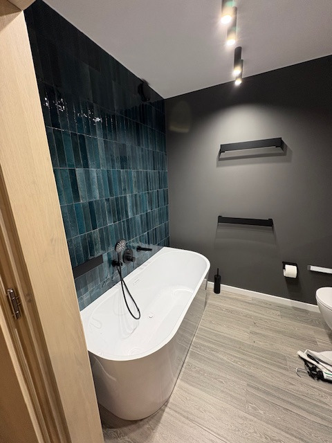 Nowoczesna łazienka z wanną wolnostojącą, turkusowymi płytkami na ścianie i szarą ścianą. Minimalistyczne półki i oświetlenie punktowe.