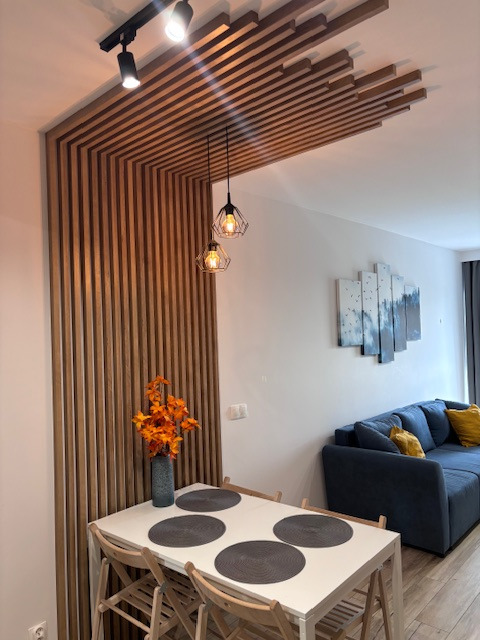 Nowoczesna aranżacja wnętrza z drewnianymi lamelami na ścianie i suficie, oświetleniem punktowym i wiszącymi lampami nad stołem jadalnym. W tle niebieska sofa.