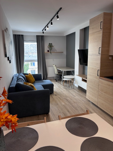 Nowoczesne wnętrze salonu z aneksem kuchennym: szara sofa narożna, drewniana podłoga, stół jadalny z okrągłymi podkładkami i oświetlenie szynowe.