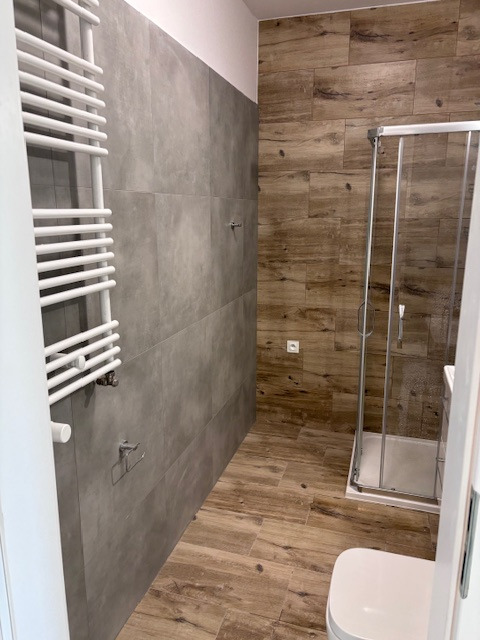 Nowoczesna łazienka z szarymi płytkami ściennymi i drewnopodobną podłogą. Widoczny grzejnik drabinkowy, kabina prysznicowa i toaleta. Minimalistyczny design.