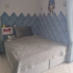 Łóżko z wezgłowiem w kształcie rombów z jasnoniebieskiej tkaniny, szary koc z geometrycznym wzorem, biała szafka nocna z zabawkami, łapacz snów na ścianie.